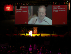 Frank Bsirske am Podium und groß auf der Bildwand.