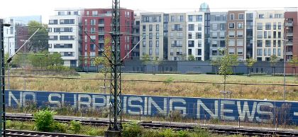 Wohnblocks hinter Bahnlinie mit Schallschutzzaun. Beschriftung: »No surprising news…«.